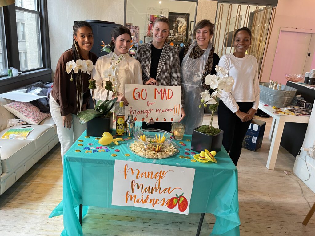 A team posing proudly behind their colorful 'Mango Mama Madness' station, showcasing their creativity in a fun and vibrant atmosphere during a team building event.