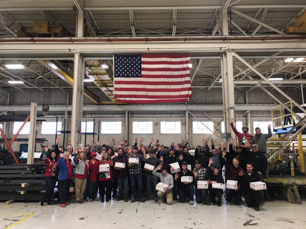 Large group of participants proudly holding care packages for military personnel, standing beneath the American flag during the Operation Military Care team building event.