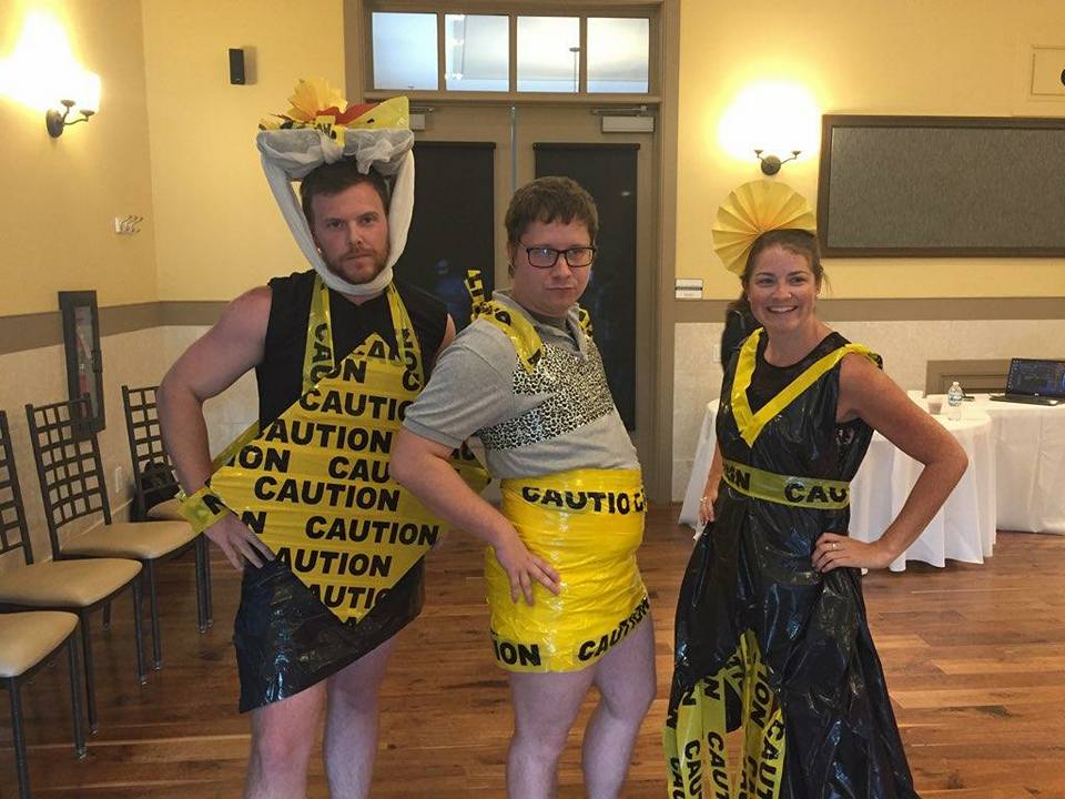 Three individuals pose confidently at a team building event, showcasing their creative outfits made from 'Caution' tape and other recyclable materials. The person on the left is wearing a black and yellow outfit, with a decorative headpiece made from caution tape. The person in the middle is also adorned in a yellow caution-tape skirt and a gray top. The individual on the right is dressed in a black and yellow dress with a matching headpiece, all made from caution tape and other materials. The mood is playful, and the group looks proud of their fashion designs.