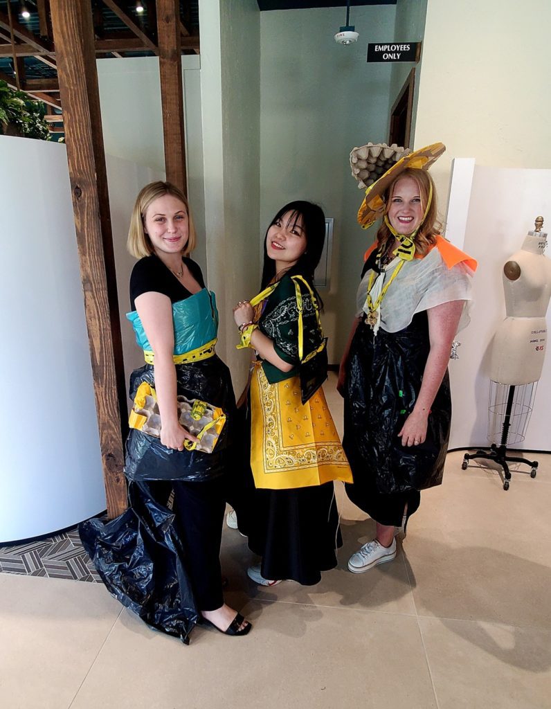 Three women standing together, smiling and posing for a photo during a team building activity. They are dressed in creative outfits made from various unconventional materials like trash bags, plastic, caution tape, and fabric. One of the women wears a large decorative hat made from a mix of recyclable materials.