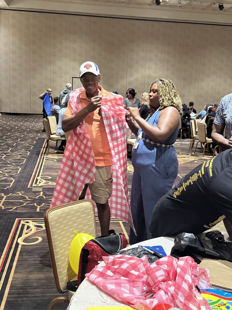 A man wearing a baseball cap is being fitted with a checkered red and white fabric draped over his shoulders, resembling a makeshift outfit. He is being assisted by a woman standing next to him, who is holding part of the fabric as she adjusts the look. They are participating in a fun team building activity in a large conference room with other groups in the background working on similar projects.