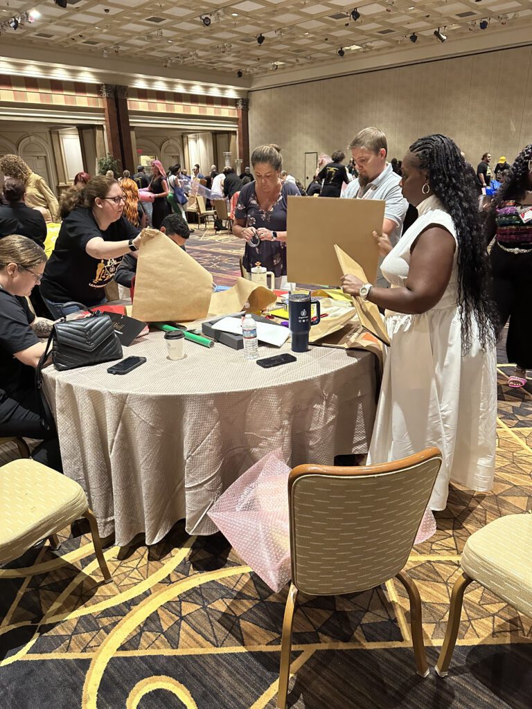 Participants at a team building event are gathered around a round table, actively engaging in a design project. They are cutting and folding pieces of cardboard and other materials as part of a creative challenge. A woman in a white dress stands next to the table, holding a piece of cardboard, while others around her are focused on their tasks. The room is filled with other groups working on similar projects.