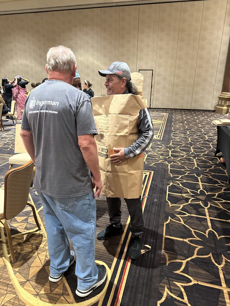 Two participants are engaged in conversation during a team building event. The man on the right is wearing an outfit made from a large brown paper bag, styled as a tunic. Both individuals appear to be enjoying the activity, and the event is clearly focused on fashion, creativity and fun.