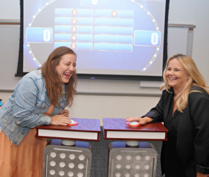 Two participants laughing and enjoying themselves while standing at game show podiums with buzzers, ready to answer a question during the Corporate Feud team building event, with a game board visible in the background.