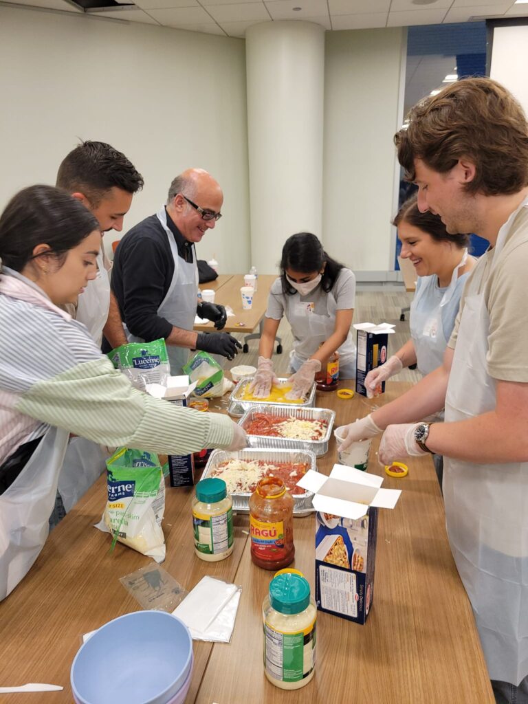 A group of six participants, wearing aprons and gloves, are gathered around a table during a team building cooking activity. They are preparing a lasagna dish using ingredients such as pasta sauce, cheese, and lasagna sheets. The group appears cheerful and collaborative as they layer ingredients into trays. Various jars, boxes, and containers are scattered on the table as they work together to complete the task.
