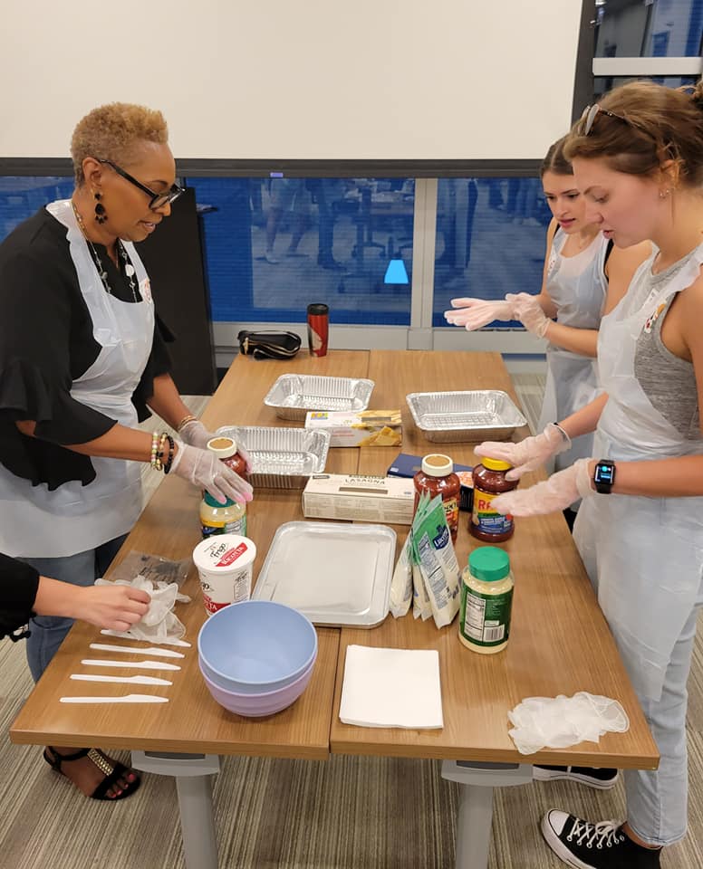 A group of four women, wearing aprons and gloves, are standing around a table during a team building cooking activity. Various ingredients, such as jars of pasta sauce, parmesan cheese, and lasagna trays, are placed on the table, along with mixing bowls and utensils. They are preparing to assemble a dish, with one woman holding a jar of sauce and the others ready to begin. The group appears focused, engaged in the process, and collaborating on the cooking task.