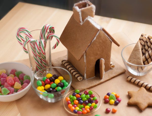 A partially constructed gingerbread house sits on a wooden table surrounded by bowls of colorful candies, including gumdrops, Skittles, and candy canes. The gingerbread house features a tall roof and chimney, with wafer sticks used as decorative accents along the base. The scene is ready for further decoration during the team building event.