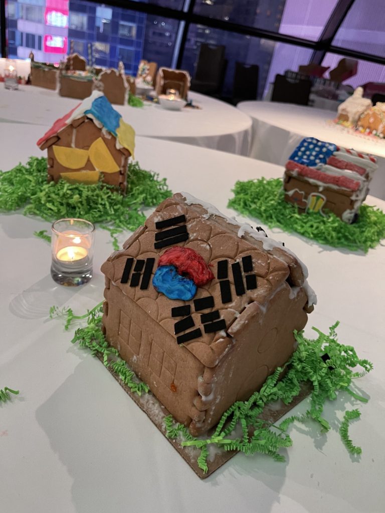 A gingerbread house decorated with icing and black licorice pieces, featuring the South Korean flag symbol in the center of the roof. The house is placed on a white table with green decorative grass around it, alongside a small lit candle. Other uniquely decorated gingerbread houses are visible in the background as part of a team building event.