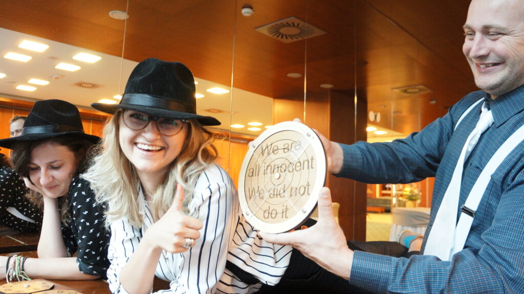 Participants in themed costumes, including hats and suspenders, holding up a clue that reads 'We are all innocent, We did not do it' during the 'Escape the Mob' team building event.