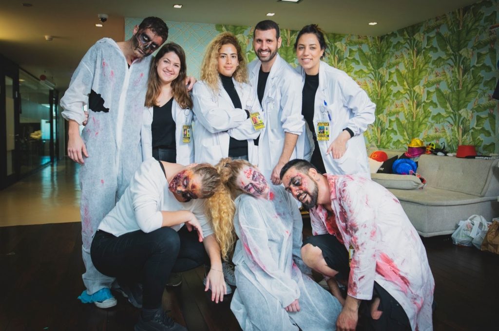 A group of people dressed in white lab coats and zombie makeup, posing together indoors. Some are wearing realistic zombie face paint and bloody costumes, while others are in scientist attire, creating a dynamic contrast between survivors and the undead in a zombie-themed team building event.