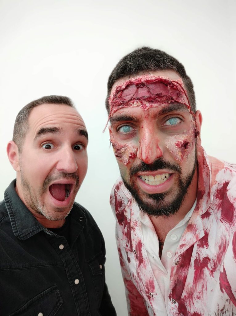 A man excitedly poses next to a participant dressed as a zombie, complete with realistic prosthetic makeup featuring bloody wounds, decaying skin, and white contact lenses, creating an eerie and convincing undead appearance during a zombie-themed team building event.